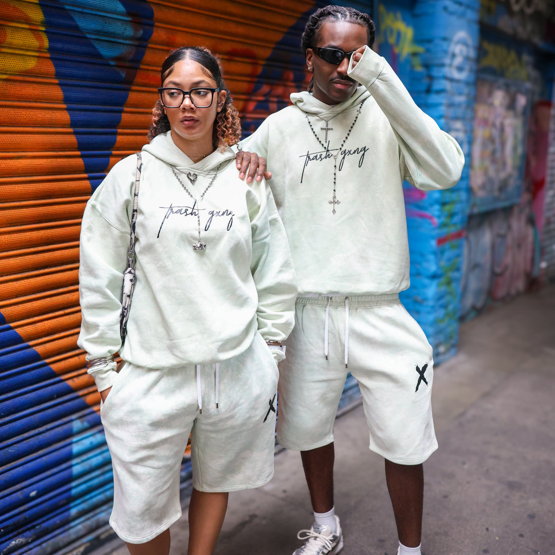 A man and a woman wearing matching teal-green streetwear 2 piece sets with 'Trash Gxng' text on the hoodies and shorts, standing in front of a colorful graffiti-covered metal shutter.