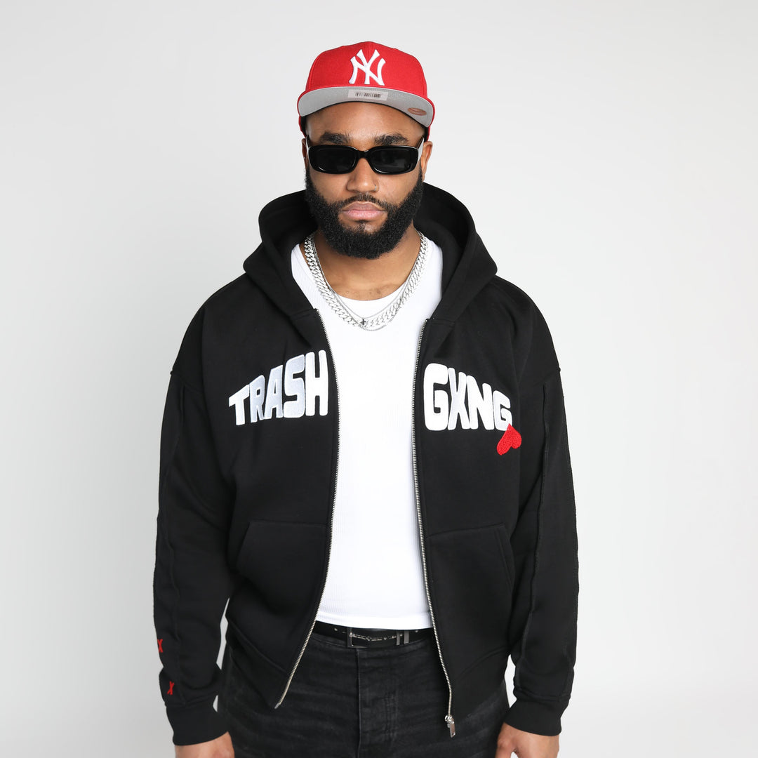 Man wearing a black hoodie with 'Trash Gxng' text, white shirt, red cap, and sunglasses on a white background