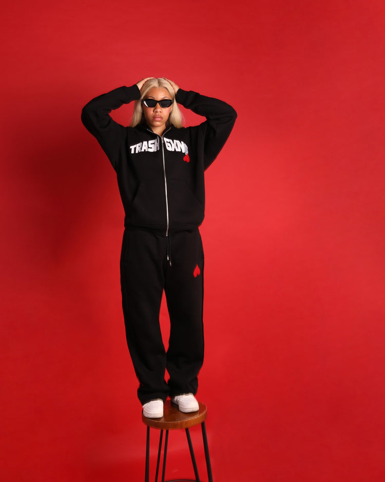 Woman wearing a black tracksuit with visible branding on a red background standing on top of a chair